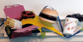 sculptures of children's fast food meal in a box including a burger, french fries in a bag, drink spilling out of a cup, and toy car